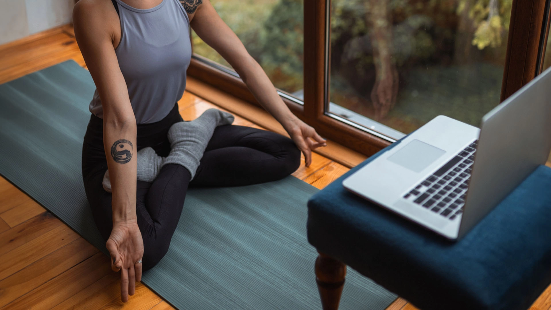Person in gray tank top and black leggings sitting cross-legged on a blue yoga mat, meditating near a window with an open laptop nearby.